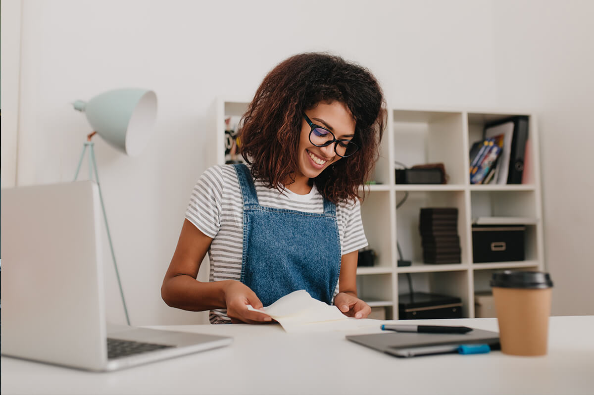 Young woman wearing glasses and blue overalls preparing papers at a desk with minimal decor