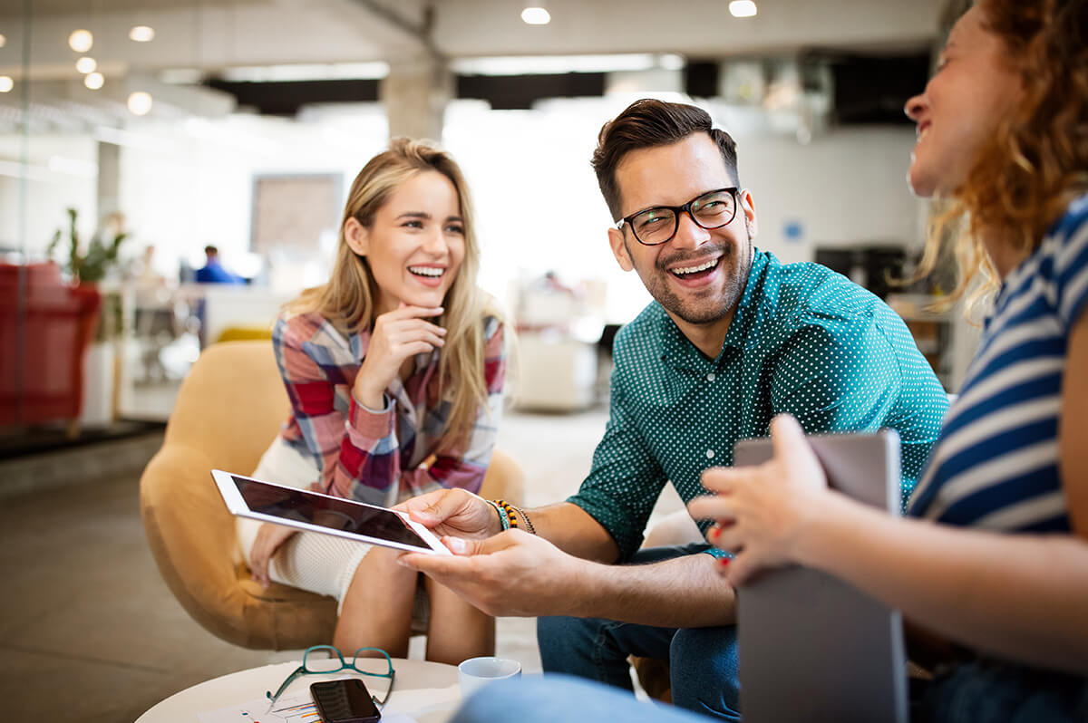 Three young professional people wearing smart casual clothing sitting together smiling, two holding tablets