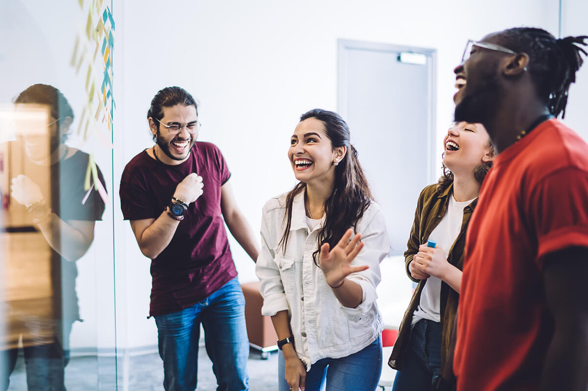 Young adults wearing smart casual clothing laughing while looking at a glass board with post-it notes