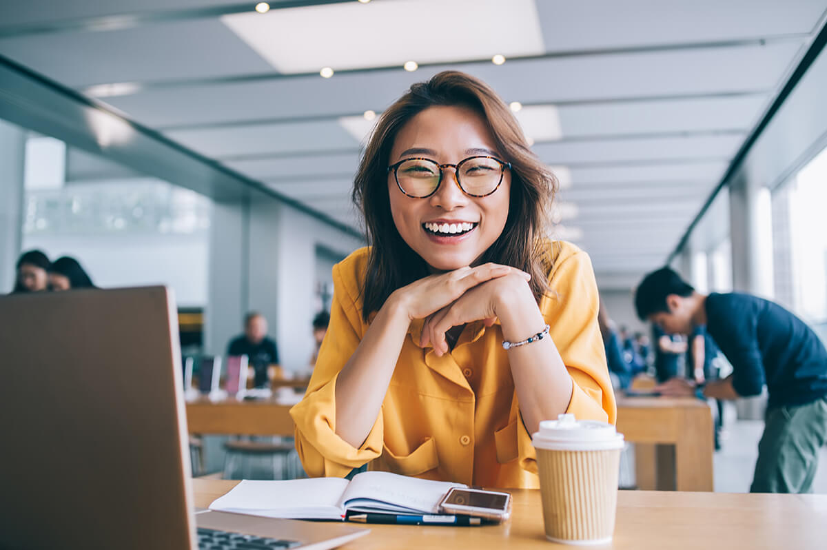 Woman with yellow blouse smiling at desk while using laptop, communal office setting behind her